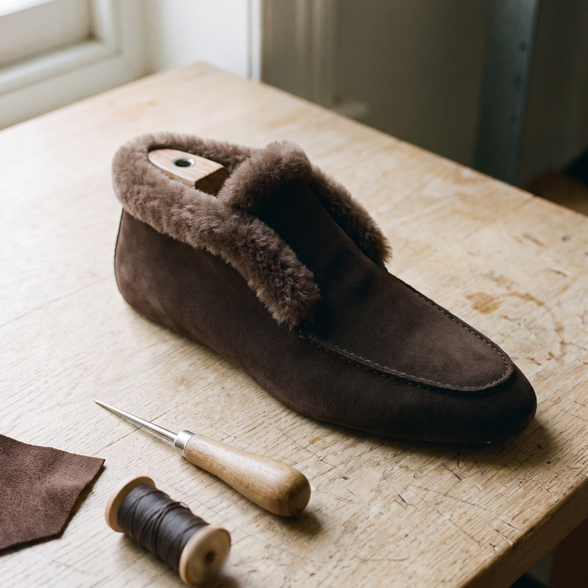 An unfinished suede loafer on a wooden last with an awl and a spool of thread on a workbench